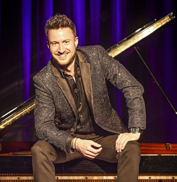 Ein Mann in einem glitzernden Anzug sitzt l&auml;chelnd vor einem Fl&uuml;gel auf einer B&uuml;hne mit violettem Hintergrund., &copy; Stadthalle Leonberg