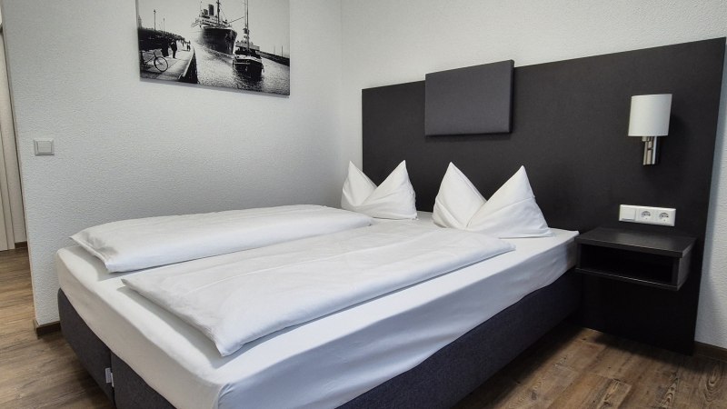 Modernes Hotelzimmer mit Doppelbett, weißen Bettdecken, Nachttisch und Wandbild eines Schiffs. Helle Wände und Holzboden., © MedientechnikKeuk