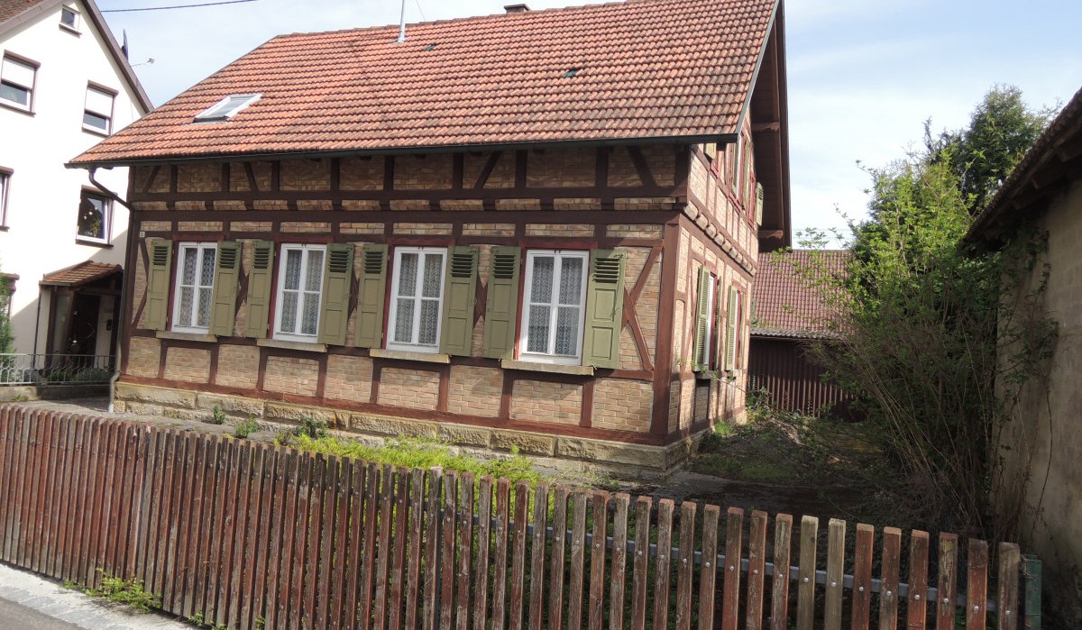 Fachwerkhaus mit roten Dachziegeln und grünen Fensterläden, umgeben von einem Holzzaun. Im Hintergrund sind weitere Gebäude sichtbar., © Land der 1000 Hügel - Kraichgau-Stromberg Fachwerkhaus mit roten Dachziegeln und grünen Fensterläden, umgeben von einem Holzzaun. Im Hintergrund sind weitere Gebäude sichtbar., © Land der 1000 Hügel - Kraichgau-Stromberg