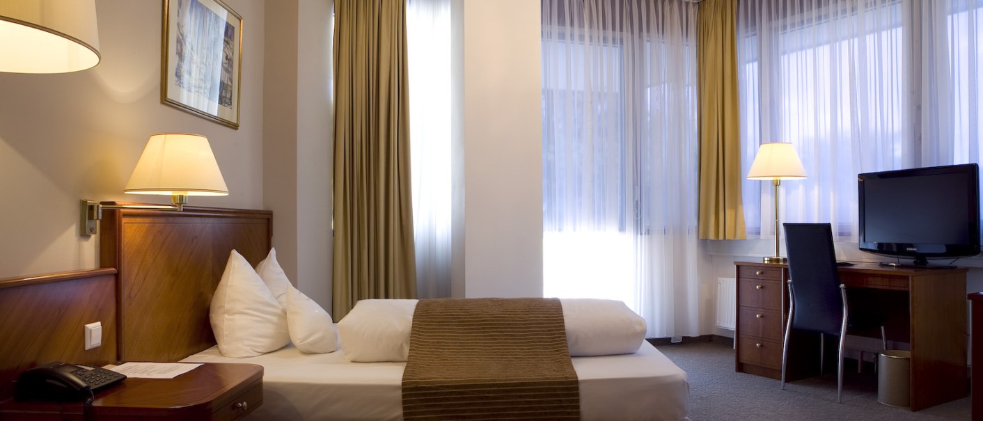 Ein elegantes Einzelzimmer mit Bett, Schreibtisch, Stuhl und Fernseher. Vorhänge und ein Kronleuchter verleihen dem Raum eine luxuriöse Atmosphäre., © Ross Messehotel Ein elegantes Einzelzimmer mit Bett, Schreibtisch, Stuhl und Fernseher. Vorhänge und ein Kronleuchter verleihen dem Raum eine luxuriöse Atmosphäre., © Ross Messehotel