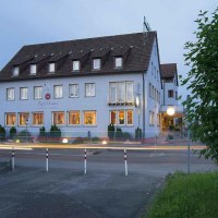 Ein Hotel mit beleuchteten Fenstern bei Dämmerung. Autos fahren vorbei, was Lichtspuren erzeugt. Das Gebäude hat ein Satteldach und mehrere Etagen., © Nödinger Gastro GmbH Ein Hotel mit beleuchteten Fenstern bei Dämmerung. Autos fahren vorbei, was Lichtspuren erzeugt. Das Gebäude hat ein Satteldach und mehrere Etagen., © Nödinger Gastro GmbH