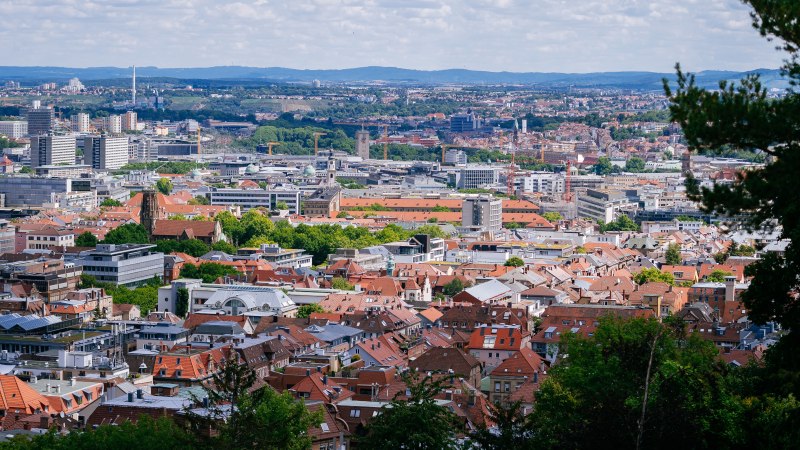 Panoramablick auf eine Stadt mit roten Dächern und modernen Gebäuden, umgeben von grünen Hügeln und bewölktem Himmel., © Thomas Niedermüller