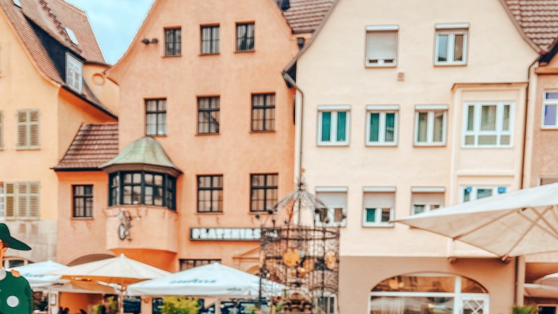 Der Hans-im-Glück-Brunnen vor historischen Gebäuden. Im Vordergrund sind Sonnenschirme und Figuren zu sehen. Die Häuser haben rote Dächer und viele Fenster., © SMG, takemyhearteverywhere Der Hans-im-Glück-Brunnen vor historischen Gebäuden. Im Vordergrund sind Sonnenschirme und Figuren zu sehen. Die Häuser haben rote Dächer und viele Fenster., © SMG, takemyhearteverywhere