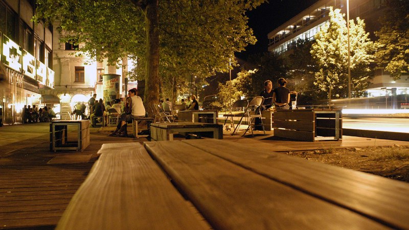 Nachtaufnahme der Theodor-Heuss-Straße in Stuttgart. Menschen sitzen auf Holzbänken unter Bäumen, umgeben von beleuchteten Gebäuden und Straßenlaternen., © Stuttgart-Marketing GmbH, Marc Gilardone