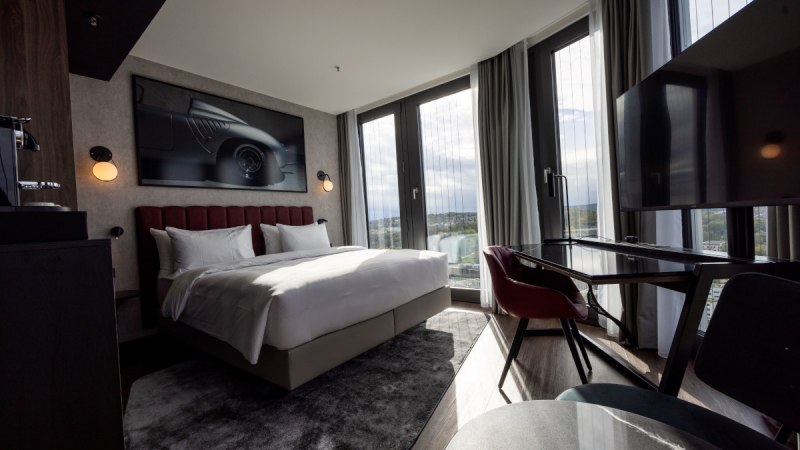 Modernes Eckzimmer mit großem Bett, Schreibtisch, Panoramafenstern und stilvollem Dekor. Ein Bild eines Autos hängt an der Wand., © Radisson Hotel Group