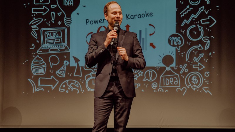 Ein Mann im Anzug spricht auf einer B&uuml;hne vor einer PowerPoint-Folie mit dem Titel 'PowerPoint Karaoke' und verschiedenen Symbolen., &copy; Stadt Leinfelden-Echterdingen, Kulturamt