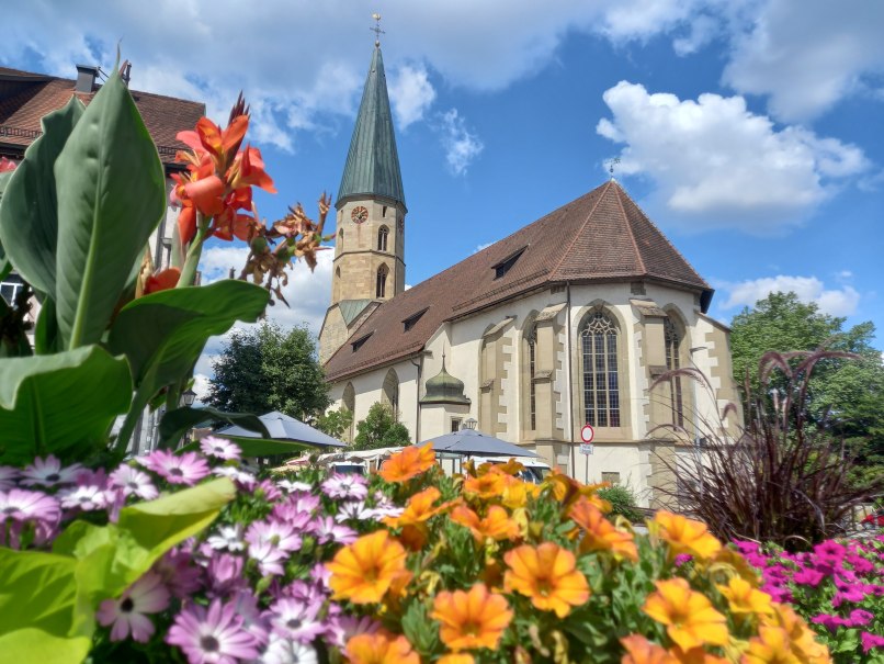 Kirche mit Kirchturm vor blauem Himmel mit weißen Wolken, davor bunte Sommerblumen, © Petra Natzkowski