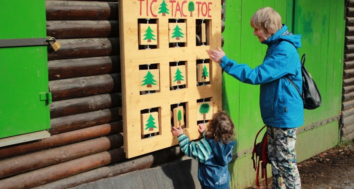 Ein Kind und eine &auml;ltere Person spielen Tic Tac Toe an einer Holzwand mit gr&uuml;nen Symbolen. Sie stehen vor einer H&uuml;tte mit gr&uuml;nen T&uuml;ren., &copy; Amt f&uuml;r Forsten Landkreis B&ouml;blingen