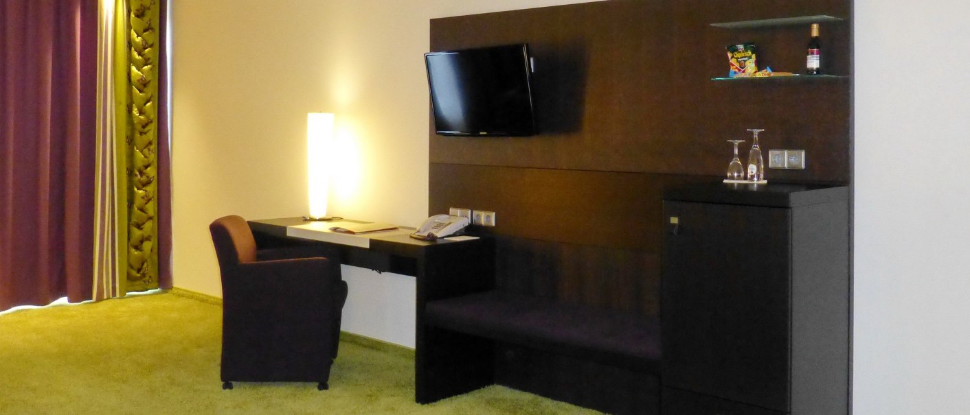 Modernes Hotelzimmer mit Schreibtisch, Sessel, Wand-TV und Minibar. Lila Vorhänge und grüner Teppich ergänzen das stilvolle Ambiente., © Hotel & Restaurant Sonne