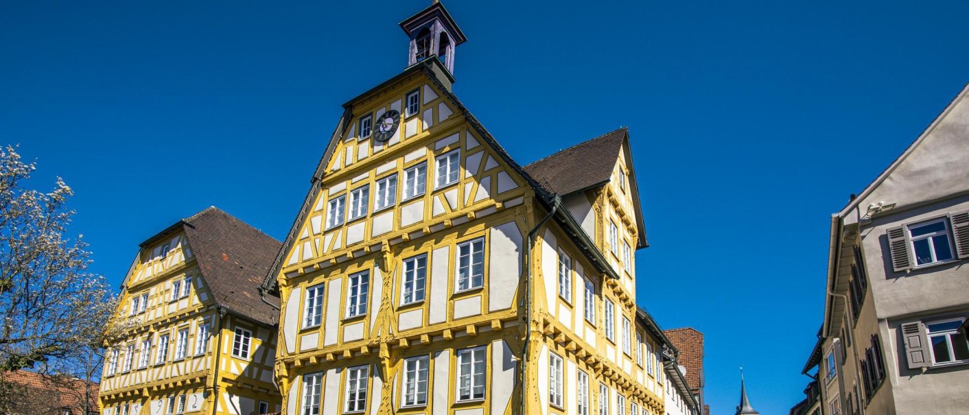 Ein historisches Fachwerkhaus in Sindelfingen, gelb und wei&szlig; gestrichen, unter klarem, blauem Himmel. Ein bl&uuml;hender Baum ist links im Bild zu sehen., &copy; SMG, Sarah Schmid