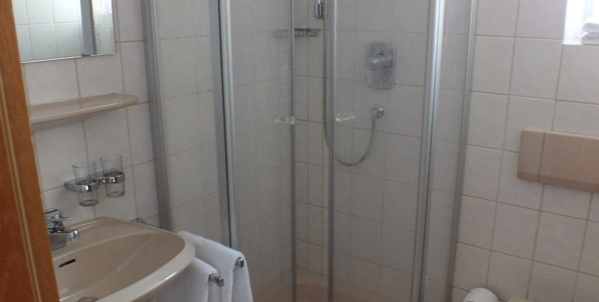 Kleines Badezimmer mit einer Eckdusche, einem Waschbecken mit Handtuch und einer Toilette. Weiße Fliesen und ein kleines Fenster sind sichtbar., © Gasthof zum Bären