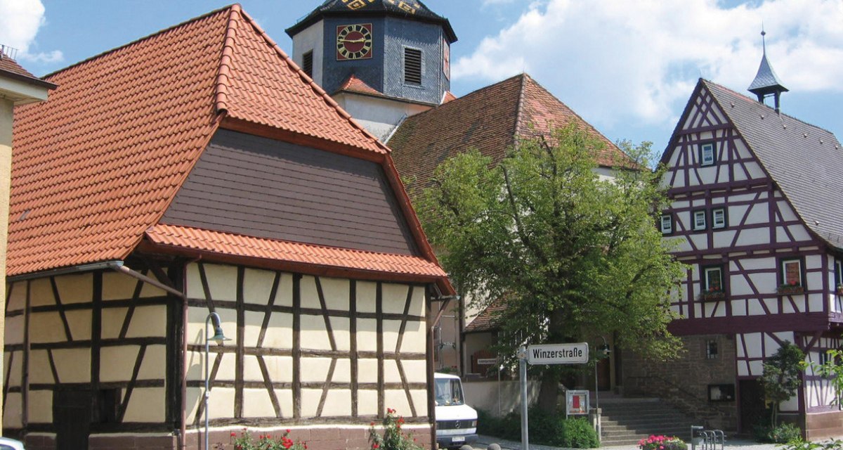 Fachwerkhäuser in Gündelbachs Ortsmitte, mit einem Kirchturm im Hintergrund. Ein Straßenschild zeigt 'Winzerstraße'. Sonniger Tag mit blauem Himmel., © Land der 1000 Hügel - Kraichgau-Stromberg Fachwerkhäuser in Gündelbachs Ortsmitte, mit einem Kirchturm im Hintergrund. Ein Straßenschild zeigt 'Winzerstraße'. Sonniger Tag mit blauem Himmel., © Land der 1000 Hügel - Kraichgau-Stromberg