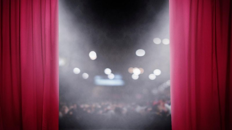 Ein roter Vorhang öffnet sich und gibt den Blick auf eine Bühne frei. Im Hintergrund ist ein unscharfes Publikum zu sehen., © AdobeStock 242383041 alphaspirit Ein roter Vorhang öffnet sich und gibt den Blick auf eine Bühne frei. Im Hintergrund ist ein unscharfes Publikum zu sehen., © AdobeStock 242383041 alphaspirit