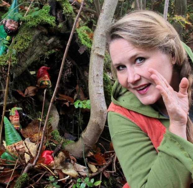 Eine Frau im Wald fl&uuml;stert zu kleinen Zwergenfiguren, die zwischen B&auml;umen und Moos versteckt sind. Sie tr&auml;gt gr&uuml;ne und orange Kleidung., &copy; Copyright: Stefanie Keller