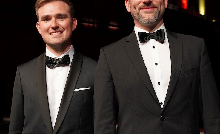 Zwei M&auml;nner in eleganten Anz&uuml;gen stehen auf einer B&uuml;hne. Der Hintergrund ist in warmen Farben beleuchtet, was eine festliche Atmosph&auml;re schafft., &copy; Touristik Bad Wildbad GmbH