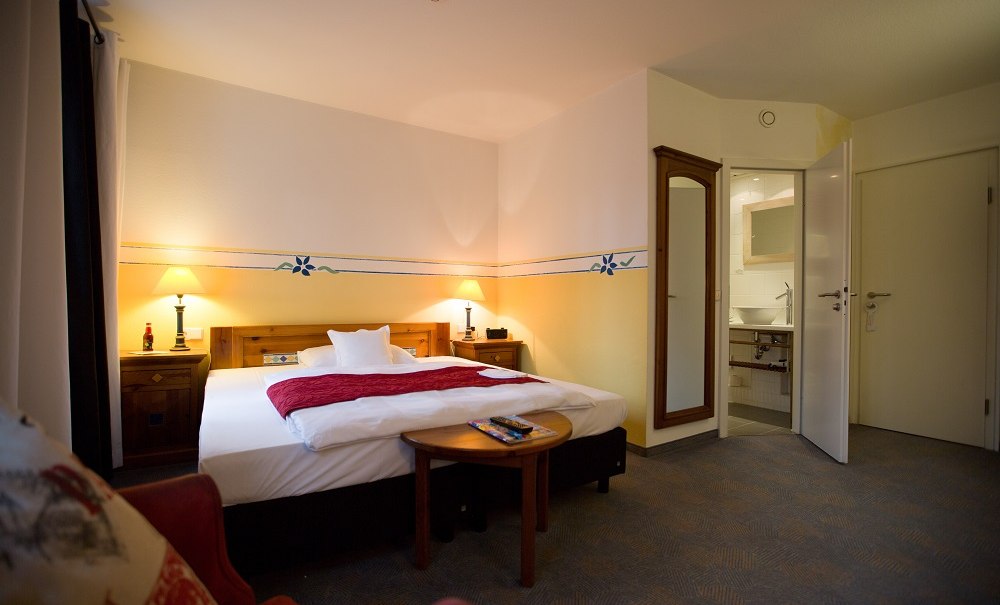Gem&uuml;tliches Hotelzimmer mit gro&szlig;em Bett, roten Akzenten, Nachttischen, Lampen und offenem Badezimmer. Ein Spiegel h&auml;ngt an der Wand., &copy; Hotel B&uuml;rkle