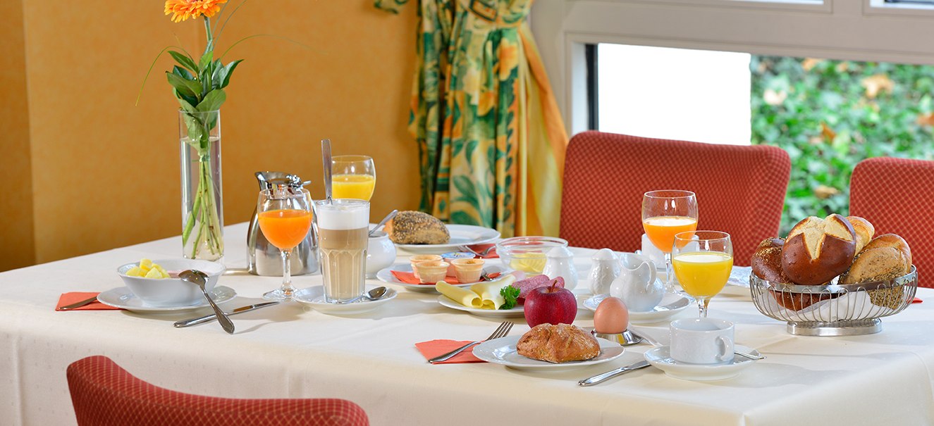 Ein Fr&uuml;hst&uuml;ckstisch mit Br&ouml;tchen, Saft, Kaffee, einem Ei, Obst und einer Blume in einer Vase, umgeben von roten St&uuml;hlen., &copy; Amber Hotel Leonberg / Stuttgart
