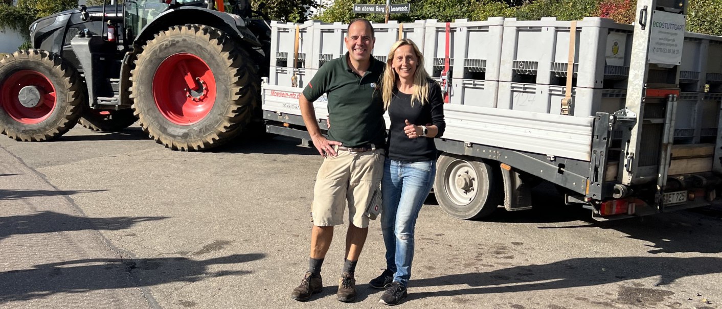 Zwei Personen posieren vor einem Traktor mit Anhänger voller Kisten. Sie lächeln in die Kamera. Im Hintergrund sind Bäume und ein Straßenschild zu sehen., © weinvomhaigst