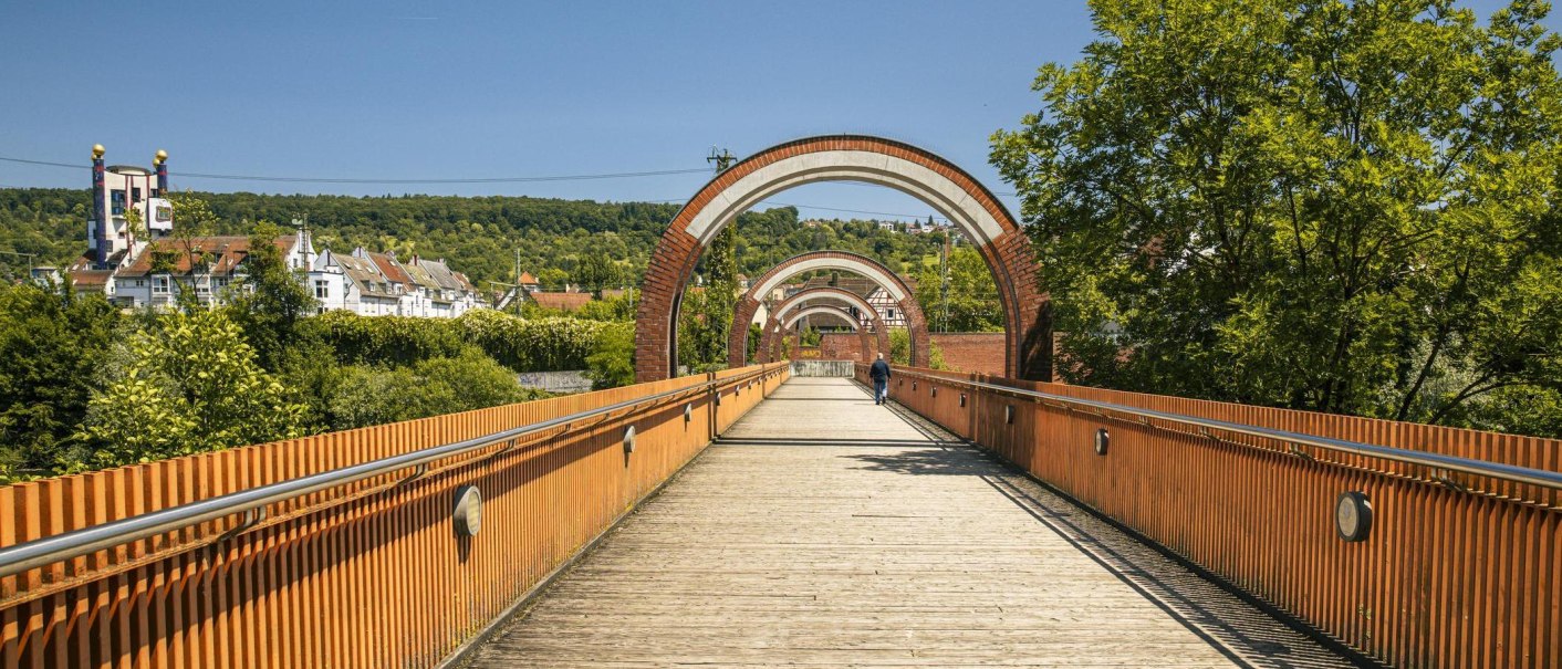 Ein Holzsteg in Plochingen mit mehreren Bögen führt durch eine grüne Landschaft. Im Hintergrund sind Gebäude und Bäume zu sehen., © SMG, Sarah Schmid Ein Holzsteg in Plochingen mit mehreren Bögen führt durch eine grüne Landschaft. Im Hintergrund sind Gebäude und Bäume zu sehen., © SMG, Sarah Schmid