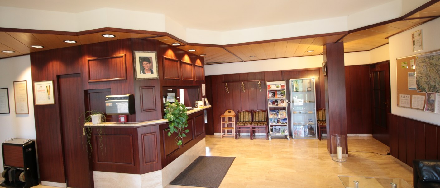 Eine Empfangshalle mit Holzwänden, einer Rezeption, Pflanzen und einem Sitzbereich. An der Wand hängen Bilder und Zertifikate., © TOMAS Eine Empfangshalle mit Holzwänden, einer Rezeption, Pflanzen und einem Sitzbereich. An der Wand hängen Bilder und Zertifikate., © TOMAS