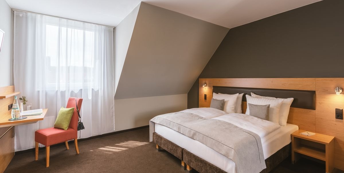 Modernes Hotelzimmer mit Doppelbett, Schreibtisch und Stuhl. Helle Vorhänge lassen Tageslicht herein. Dezente Farben und gemütliche Atmosphäre., © Novum Hospitality