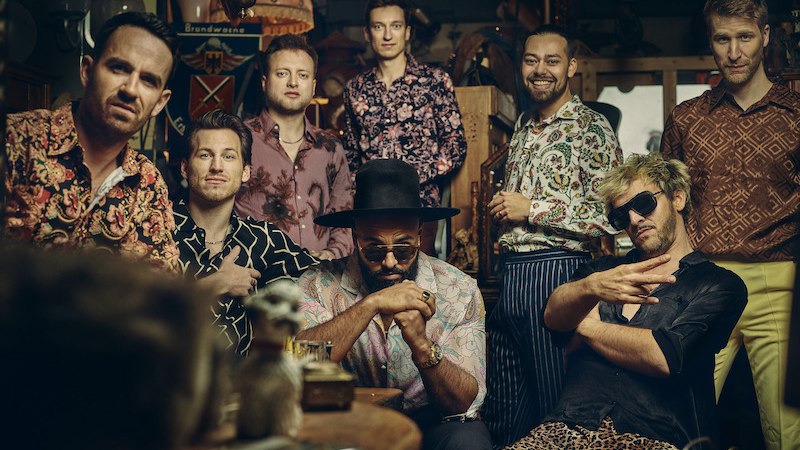 Acht Männer in bunter Kleidung posieren in einem dekorierten Raum. Einer trägt einen Hut und Sonnenbrille, andere lächeln oder schauen ernst., © Kulturhaus Schwanen Acht Männer in bunter Kleidung posieren in einem dekorierten Raum. Einer trägt einen Hut und Sonnenbrille, andere lächeln oder schauen ernst., © Kulturhaus Schwanen