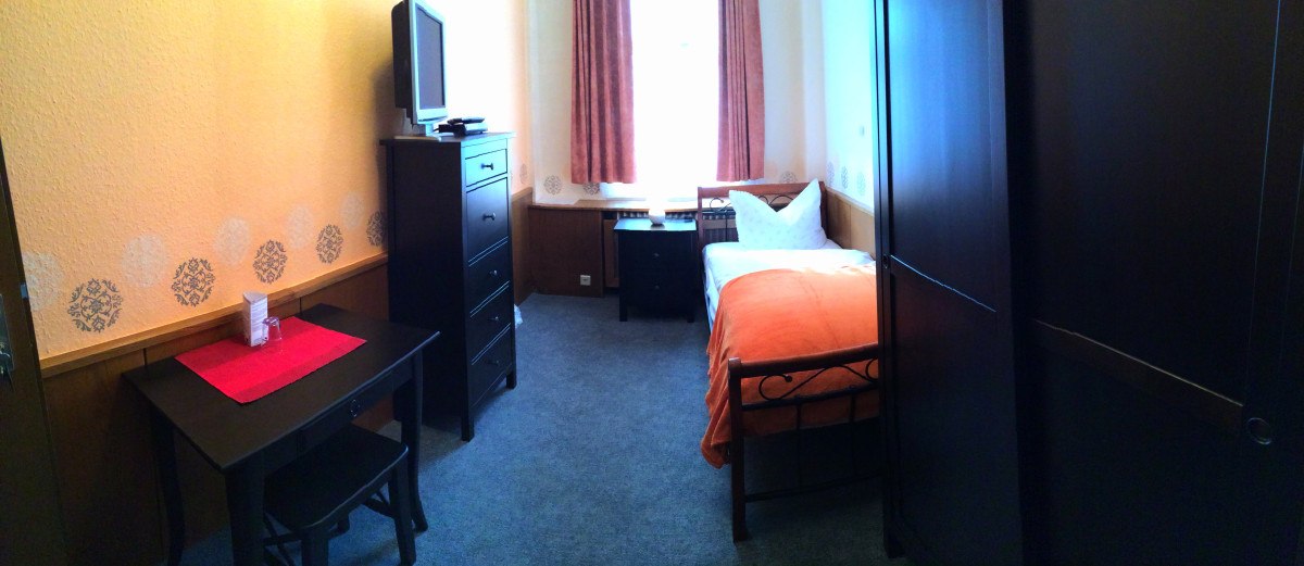 Kleines Hotelzimmer mit Einzelbett, orangefarbener Decke, Fernseher auf Kommode, Tisch mit rotem Läufer und dunklem Schrank. Kleines Hotelzimmer mit Einzelbett, orangefarbener Decke, Fernseher auf Kommode, Tisch mit rotem Läufer und dunklem Schrank.