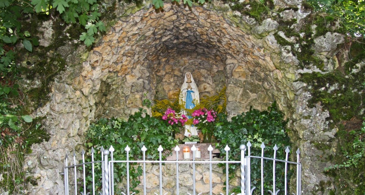 Eine Steingrotte mit einer Marienstatue, umgeben von Blumen und Pflanzen. Ein Metallzaun schützt die Grotte., © Foto: Hans-Werner Kalb Eine Steingrotte mit einer Marienstatue, umgeben von Blumen und Pflanzen. Ein Metallzaun schützt die Grotte., © Foto: Hans-Werner Kalb