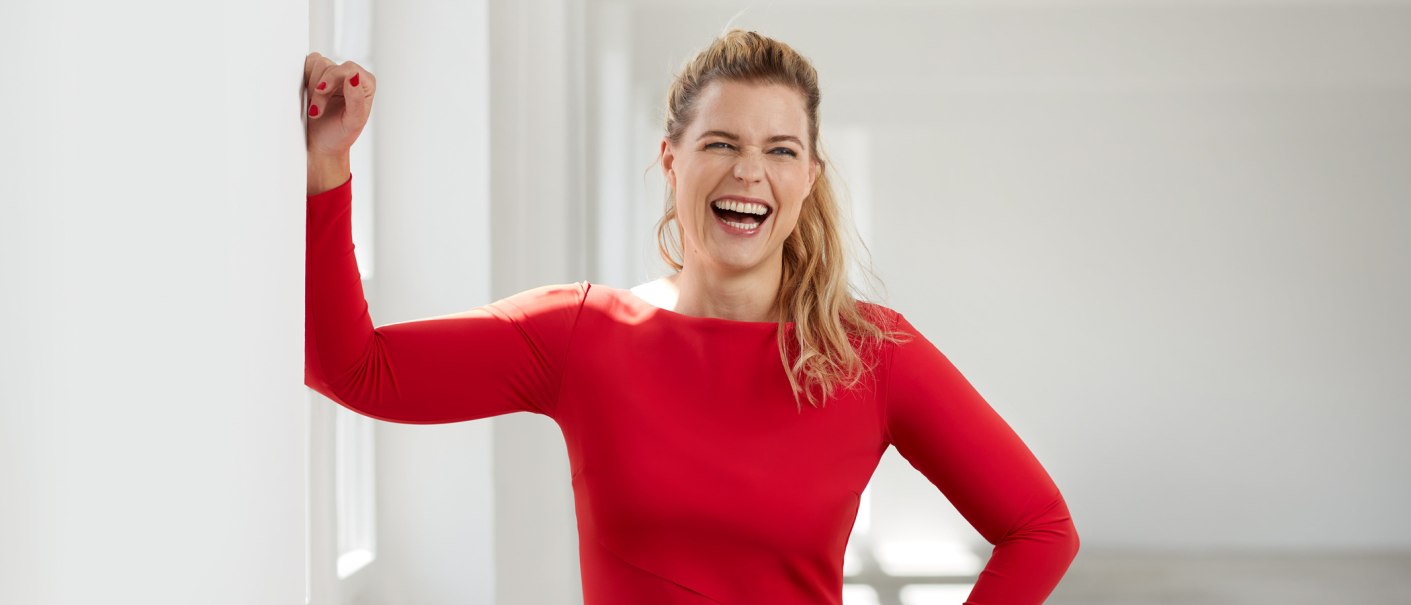 Eine Frau in einem roten Kleid lacht herzlich und lehnt sich an eine weiße Wand in einem hellen Raum., © club traube Eine Frau in einem roten Kleid lacht herzlich und lehnt sich an eine weiße Wand in einem hellen Raum., © club traube