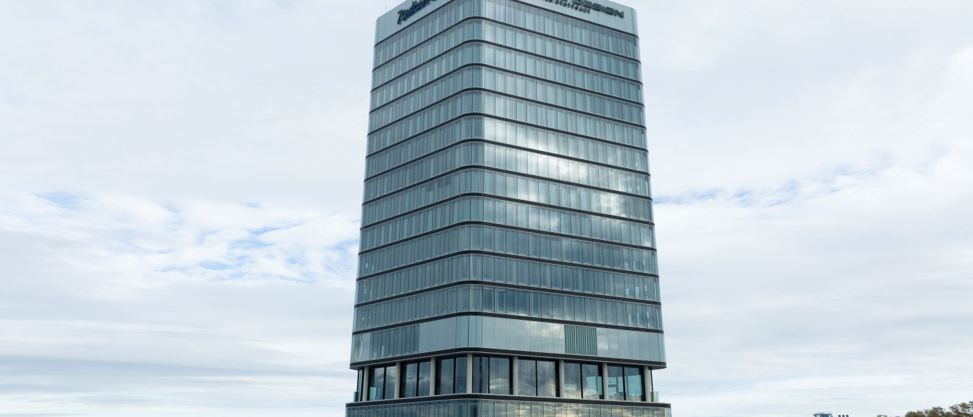 Ein modernes Hochhaus mit Glasfassade und dem Schriftzug 'Porsche Design' in einer städtischen Umgebung., © Radisson Hotel Group