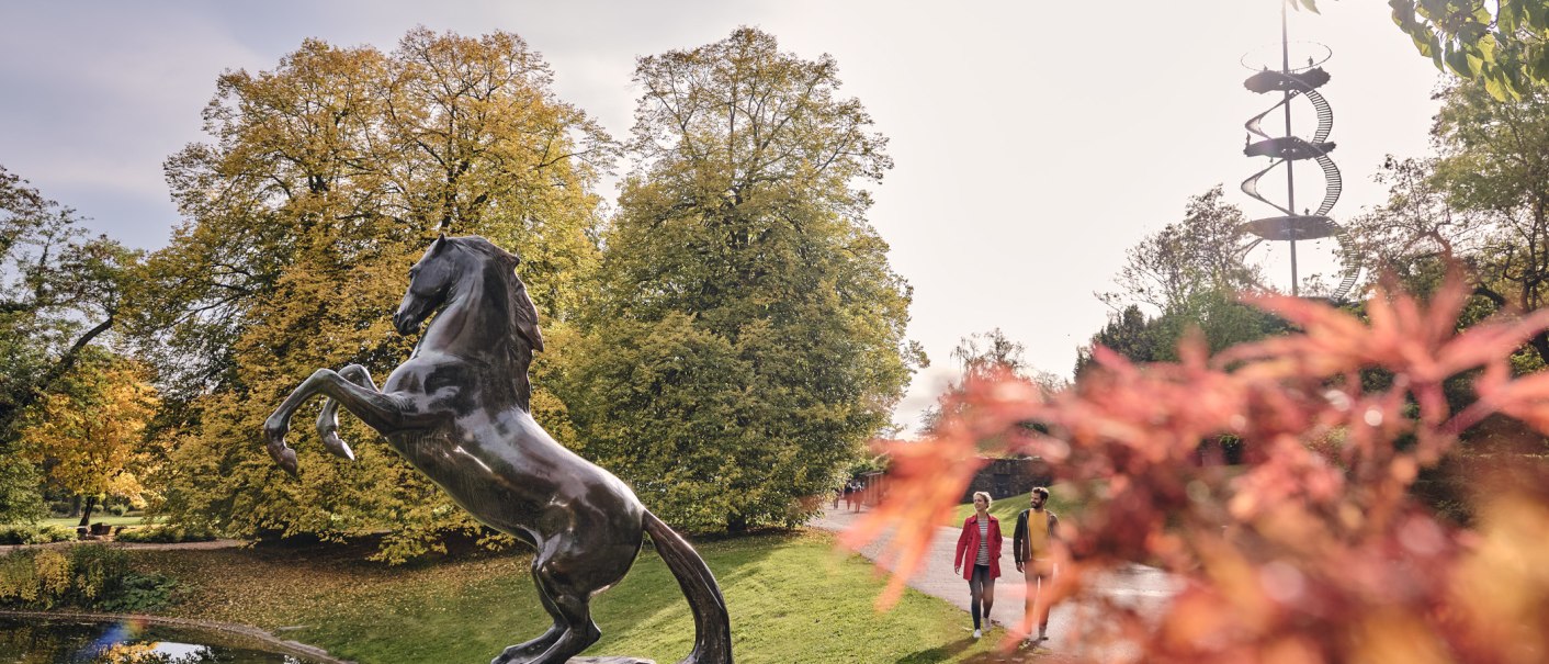 Bronzestatue eines aufsteigenden Pferdes im Höhenpark Killesberg, umgeben von herbstlichen Bäumen und einem spiralförmigen Aussichtsturm im Hintergrund., © SMG, Christoph Düpper