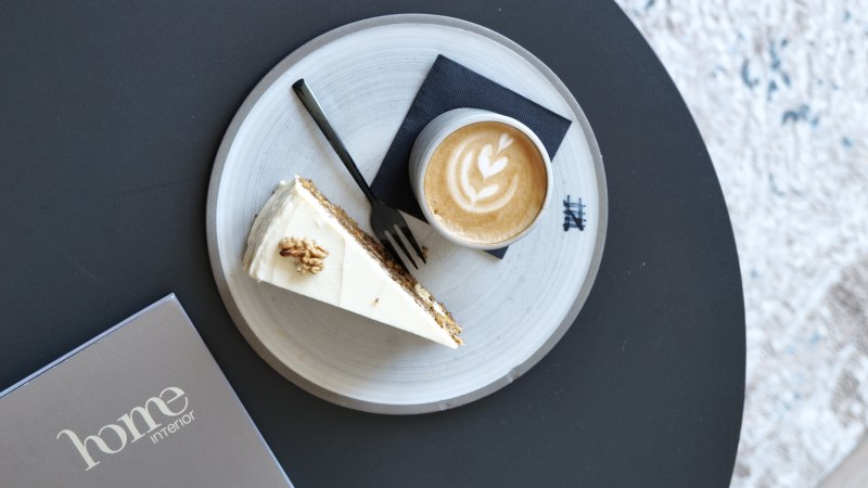Kaffee mit Latte Art und ein St&uuml;ck Kuchen auf einem Teller auf einem schwarzen Tisch, daneben eine Karte mit &bdquo;home interior&ldquo;., &copy; Prosic GmbH - TARABA