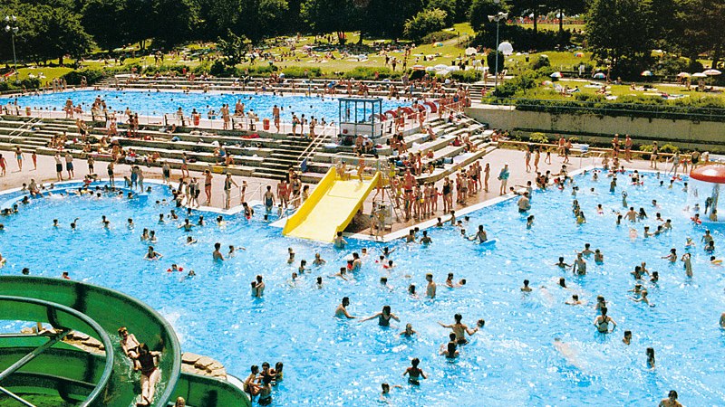 Ein belebtes Freibad mit vielen Menschen im Wasser und auf der Liegewiese. Eine gelbe Rutsche und eine gr&uuml;ne Wasserrutsche sind sichtbar., &copy; Stadt Wendlindgen am Neckar