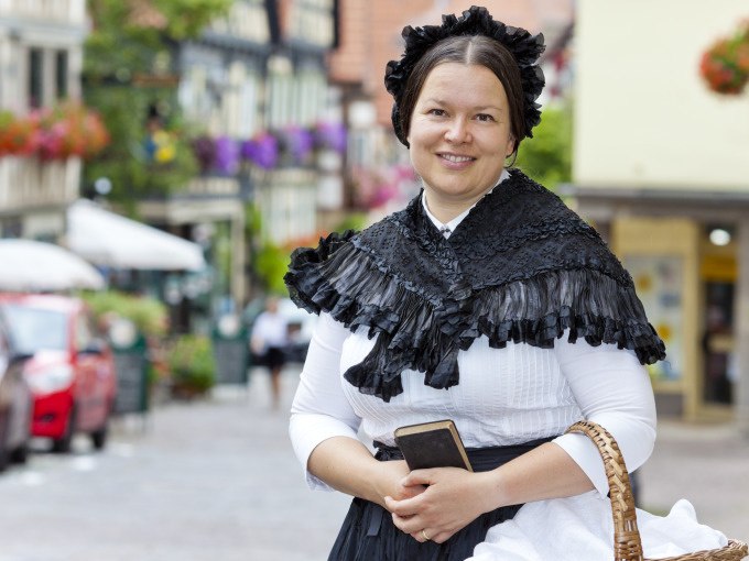 Eine Frau in historischer Kleidung des 19. Jahrhunderts steht lächelnd in einer malerischen Altstadt mit Fachwerkhäusern und Blumen., © Stadt Besigheim Eine Frau in historischer Kleidung des 19. Jahrhunderts steht lächelnd in einer malerischen Altstadt mit Fachwerkhäusern und Blumen., © Stadt Besigheim