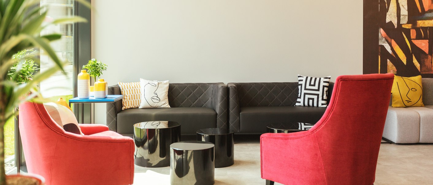 Moderne Lobby mit roten Sesseln, einem grauen Sofa und bunten Kissen. Ein blauer Tisch mit gelben Vasen und Pflanzen steht im Raum., © the niu Form - Novum Hospitality