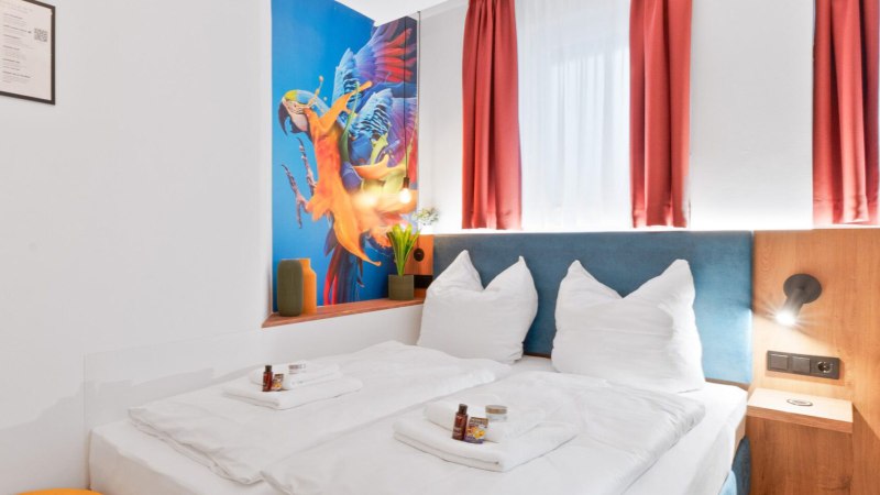 Modernes Doppelzimmer mit wei&szlig;em Bett, Papageien-Wandbild, roten Vorh&auml;ngen und gelbem Hocker. Pflegeprodukte auf dem Bett., &copy; JUNGLE N&deg;5 GmbH