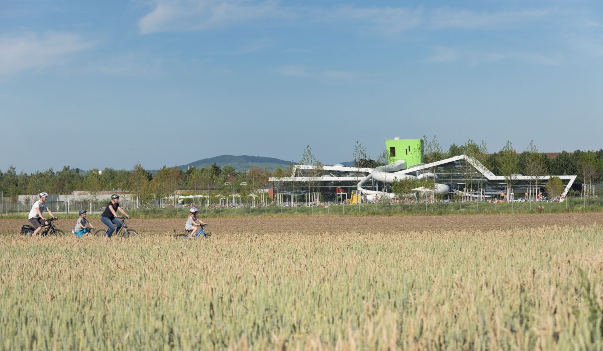 Familie radelt durch ein Feld, modernes Gebäude mit Rutschen im Hintergrund, blauer Himmel., © Stadt Fellbach Familie radelt durch ein Feld, modernes Gebäude mit Rutschen im Hintergrund, blauer Himmel., © Stadt Fellbach