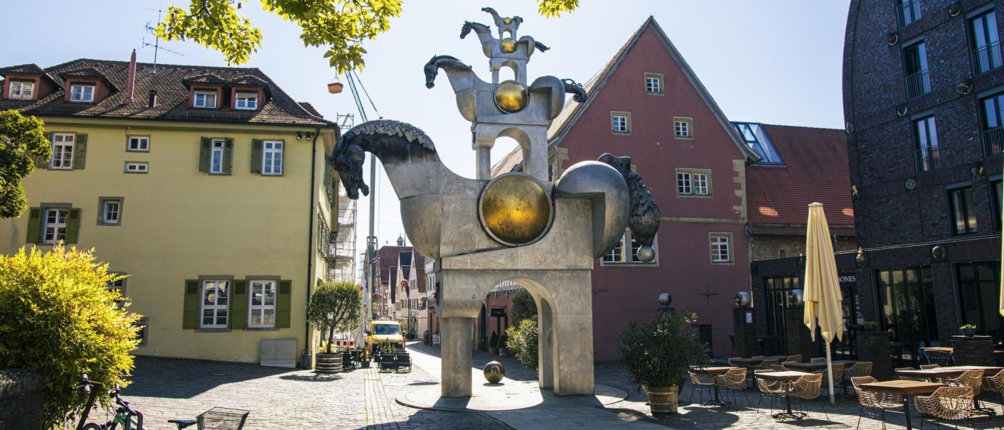 Skulptur der Grauen Pferde in Bietigheim-Bissingen, umgeben von historischen Gebäuden und einem Café mit Tischen und Stühlen im Freien., © Stuttgart Marketing GmbH, Sarah Schmid