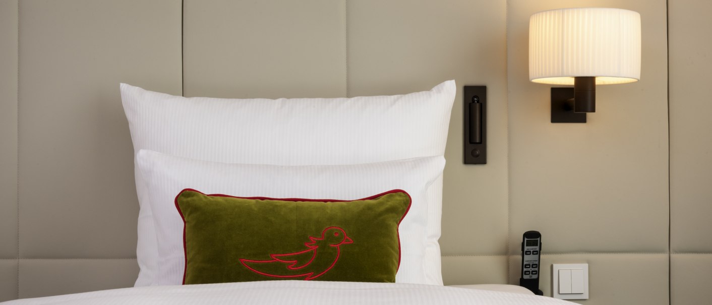 Ein Hotelzimmer mit einem weißen Bett, einem grünen Kissen mit Vogelmotiv, einer Wandlampe und einer dekorativen Tapete., © Mövenpick Hotel Stuttgart Messe & Congress Ein Hotelzimmer mit einem weißen Bett, einem grünen Kissen mit Vogelmotiv, einer Wandlampe und einer dekorativen Tapete., © Mövenpick Hotel Stuttgart Messe & Congress