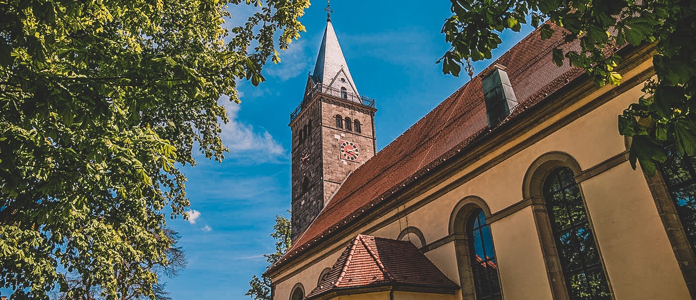 Ein Kirchturm ragt in den blauen Himmel, umgeben von grünen Bäumen. Ein historisches Gebäude mit großen Fenstern ist im Vordergrund zu sehen., © Stadt Welzheim