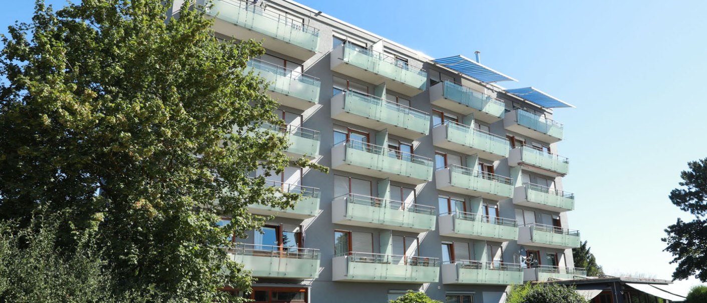 Ein modernes, mehrstöckiges Wohngebäude mit Balkonen und Glasgeländern, umgeben von Bäumen und einem klaren blauen Himmel., © TOMAS