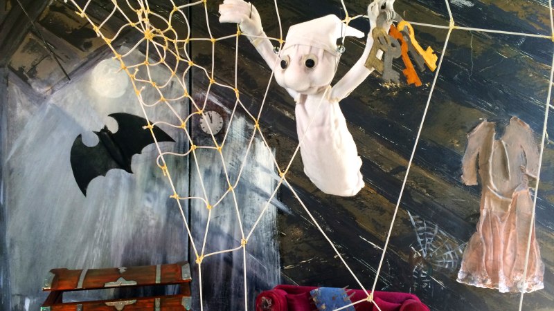Ein kleines Gespenst h&auml;ngt in einem Spinnennetz, h&auml;lt Schl&uuml;ssel. Im Hintergrund eine Fledermaus, eine offene Truhe und ein rotes Sofa., &copy; Stadtb&uuml;cherei Bad Urach