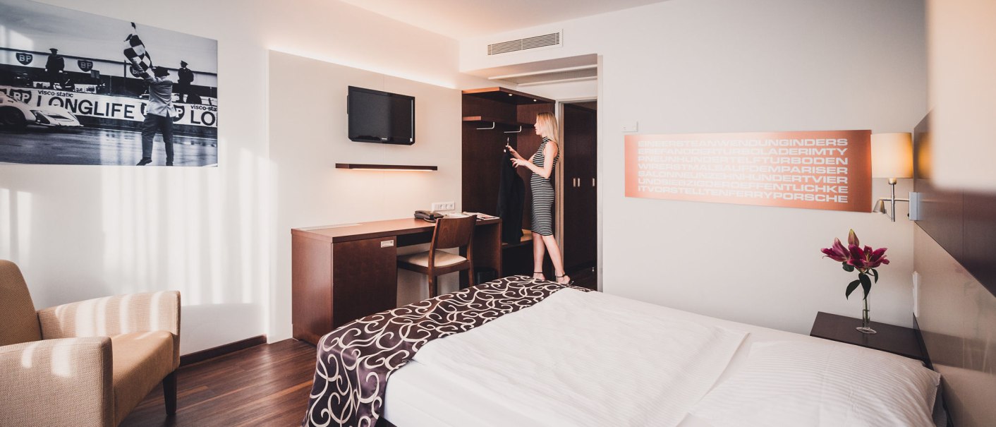 Modernes Hotelzimmer mit Doppelbett, Schreibtisch, Fernseher und Frau, die am Kleiderschrank steht. An der Wand ein Motorsportbild., © Abacco Hotels GmbH Modernes Hotelzimmer mit Doppelbett, Schreibtisch, Fernseher und Frau, die am Kleiderschrank steht. An der Wand ein Motorsportbild., © Abacco Hotels GmbH
