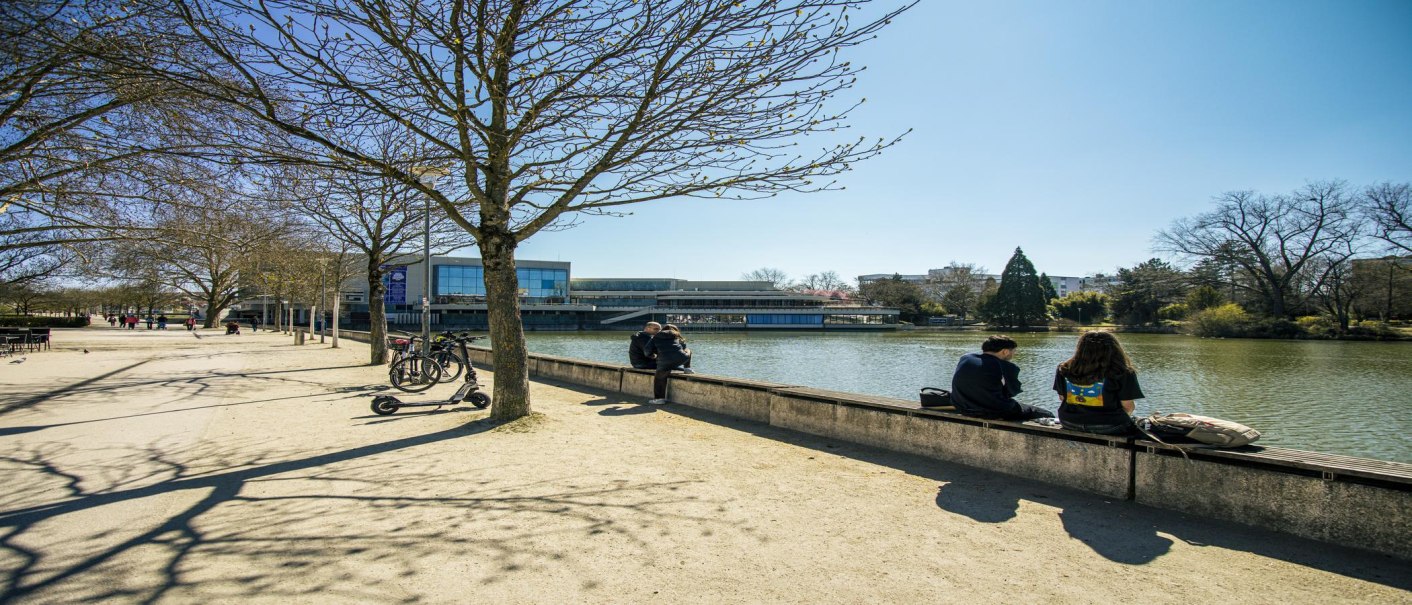 Menschen sitzen am Ufer des Unteren Sees in Böblingen. Bäume werfen Schatten auf den Weg, Fahrräder und ein Roller stehen an einem Baum., © SMG, Sarah Schmid Menschen sitzen am Ufer des Unteren Sees in Böblingen. Bäume werfen Schatten auf den Weg, Fahrräder und ein Roller stehen an einem Baum., © SMG, Sarah Schmid