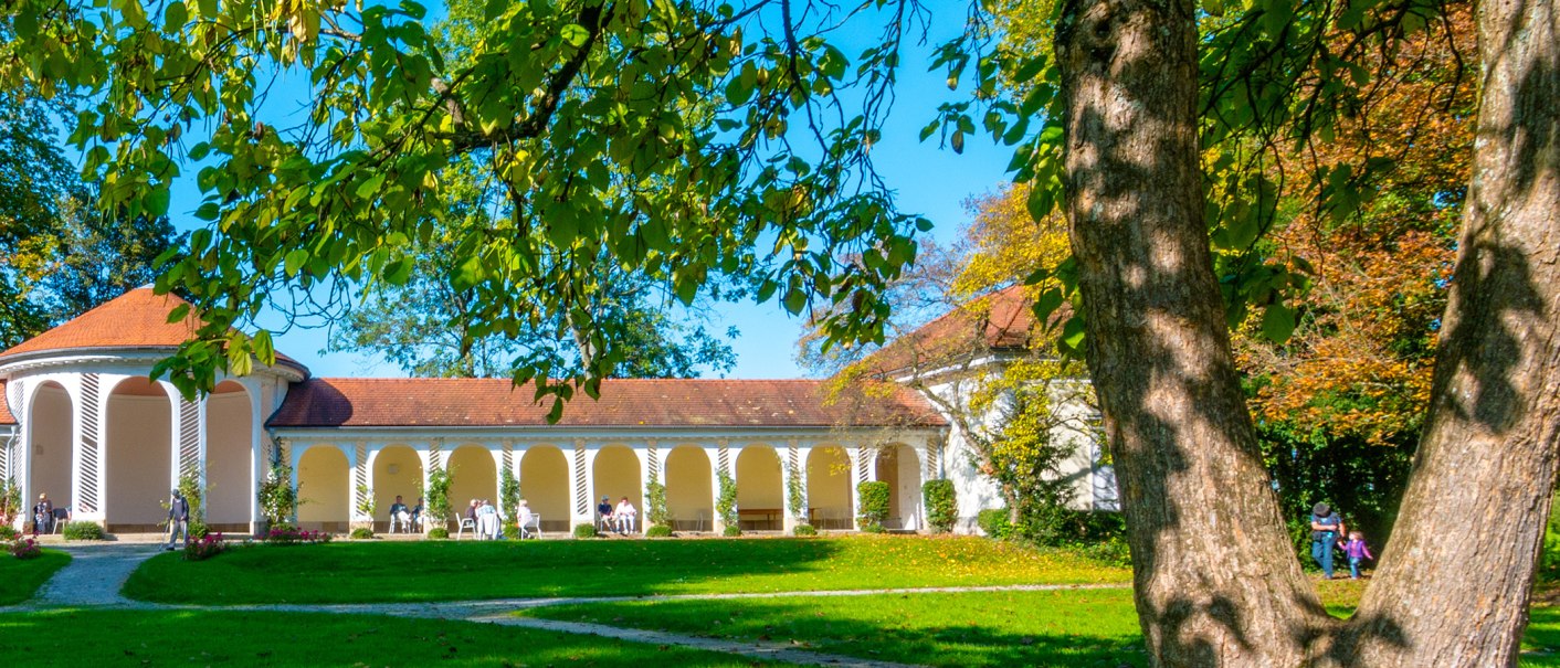 Die Wandelhalle im Kurpark Bad Boll, umgeben von grüner Wiese und Bäumen, bei sonnigem Wetter. Menschen sitzen entspannt unter den Arkaden., © Bad Boll Die Wandelhalle im Kurpark Bad Boll, umgeben von grüner Wiese und Bäumen, bei sonnigem Wetter. Menschen sitzen entspannt unter den Arkaden., © Bad Boll