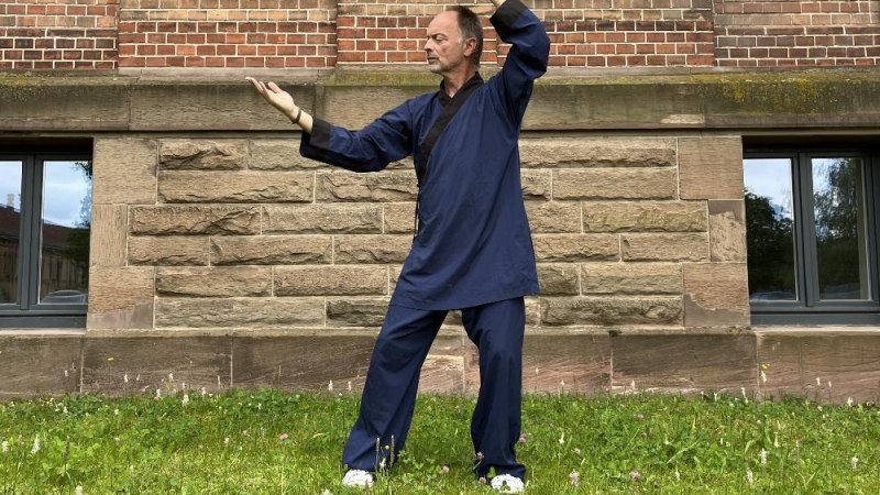 Eine Person in blauer Kleidung f&uuml;hrt eine Taiji Qigong-&Uuml;bung vor einer Backsteinmauer aus. Sie steht auf einer gr&uuml;nen Wiese., &copy; &copy; Felina Konkart