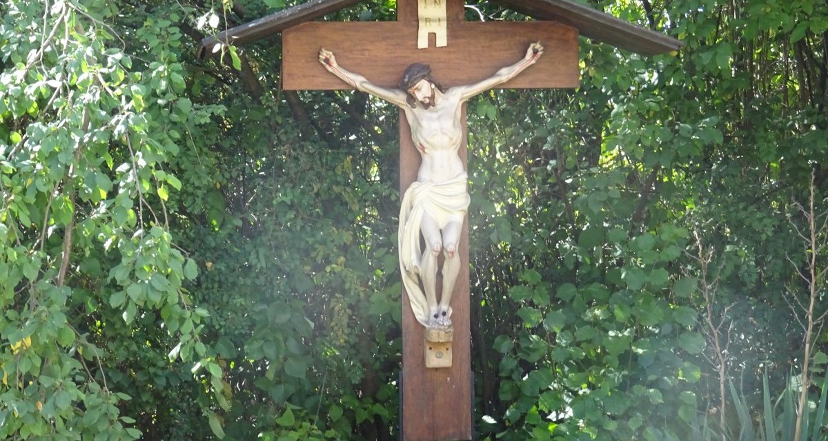Ein Kruzifix mit einer Jesusfigur steht vor dichtem gr&uuml;nen Laub. Das Kreuz ist aus Holz und hat ein kleines Dach., &copy; Foto: Cornelia Steinbach
