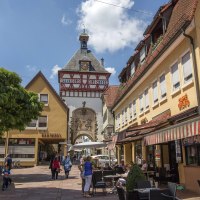 Eine belebte Altstadtstraße mit Fachwerkhaus und Uhrturm. Menschen sitzen in Straßencafés, während andere die Straße entlanggehen. Der Himmel ist blau., © Stuttgart Marketing GmbH Achim Mende