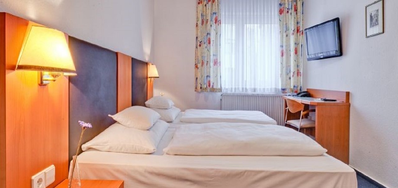 Ein gemütliches Doppelzimmer mit zwei Betten, einem Schreibtisch, einem Stuhl und einem Wandfernseher. Die Vorhänge sind bunt gemustert., © ARNOTEL GmbH Ein gemütliches Doppelzimmer mit zwei Betten, einem Schreibtisch, einem Stuhl und einem Wandfernseher. Die Vorhänge sind bunt gemustert., © ARNOTEL GmbH