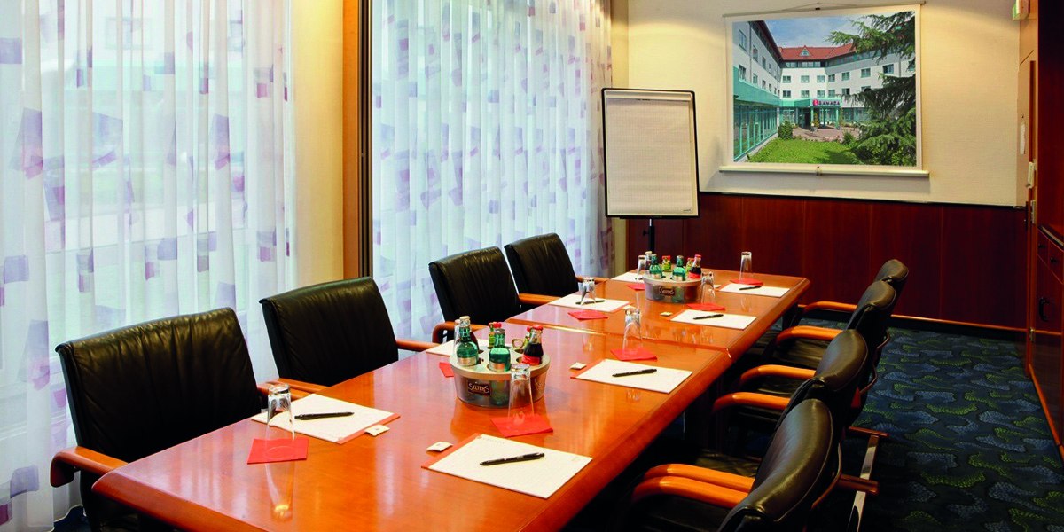 Seminarraum mit langem Tisch, Stühlen, Schreibmaterialien und Getränken. Ein Flipchart und ein Bild an der Wand sind zu sehen., © © H-Hotels.com